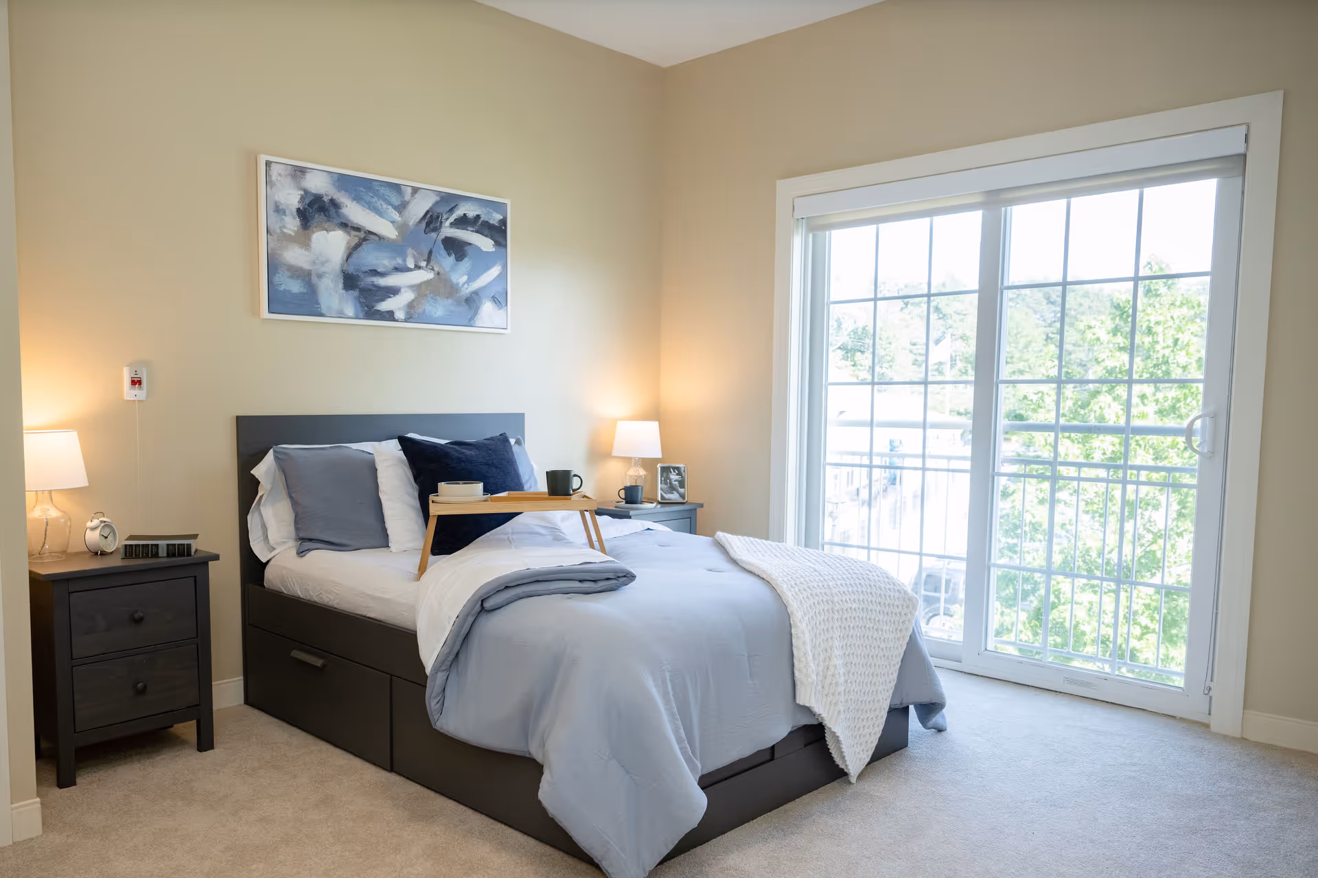 A bright bedroom with a neatly made bed featuring light blue and white bedding, two bedside tables each with a lamp, a small tray with a cup on the bed, a large window with sliding glass doors letting in natural light, and a framed abstract painting on the wall above the bed.