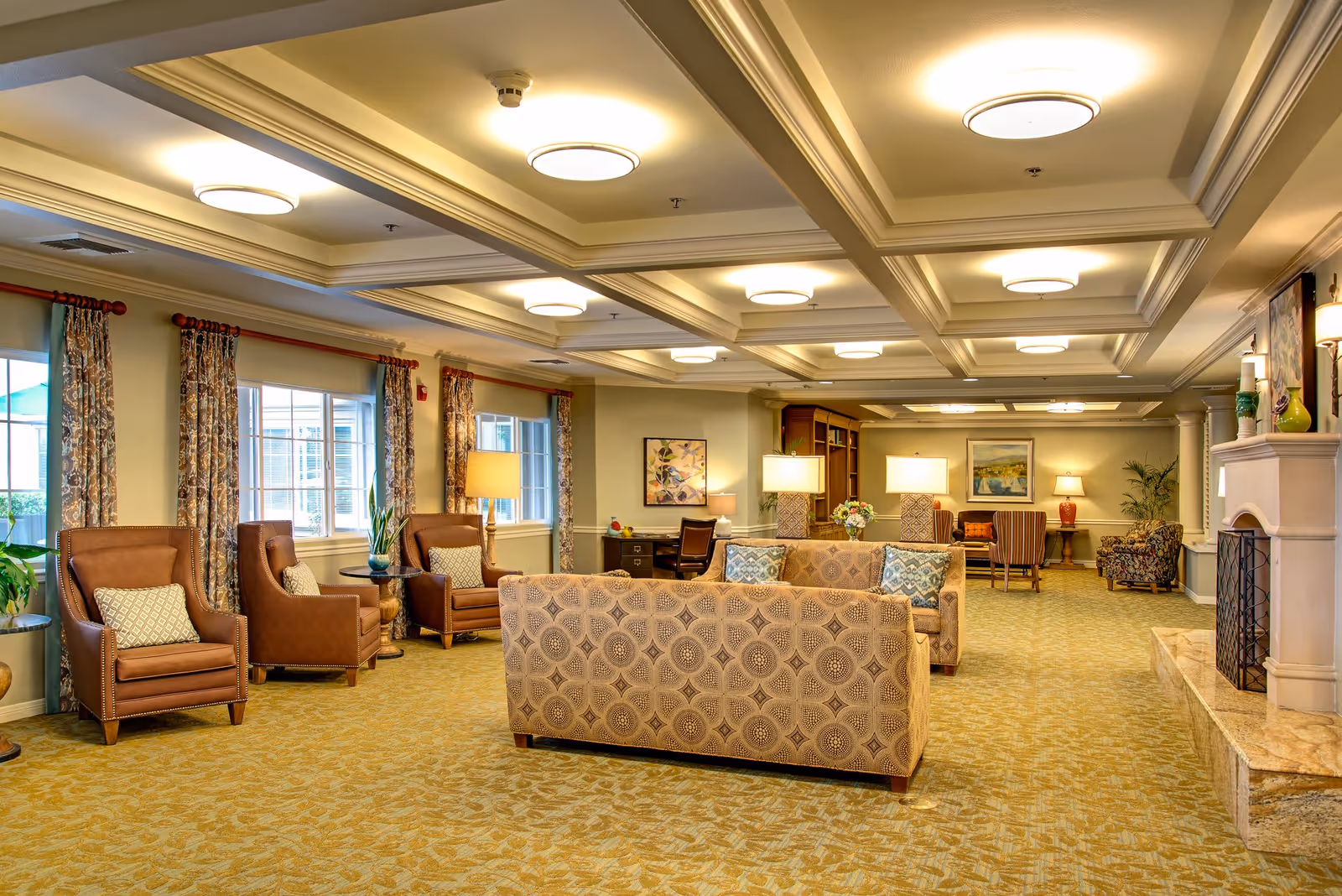 A spacious and well-lit living room area in a senior living facility with patterned sofas, leather armchairs, multiple table lamps, large windows with floral curtains, a fireplace, and decorative artwork on the walls.