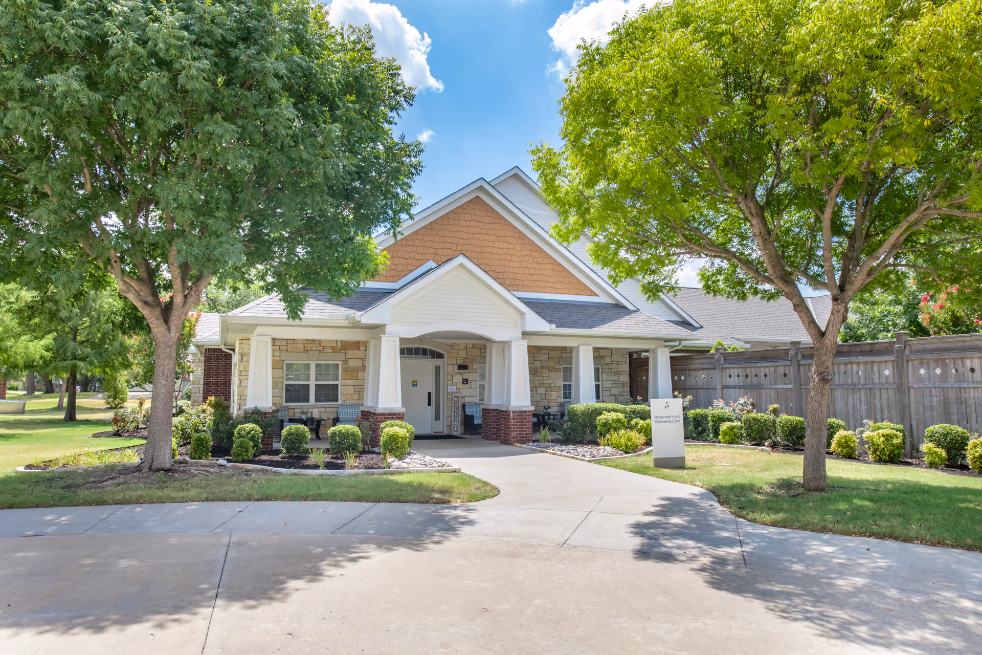 Front exterior view of a single-story building with a covered entrance, surrounded by green trees and shrubs under a blue sky with some clouds.