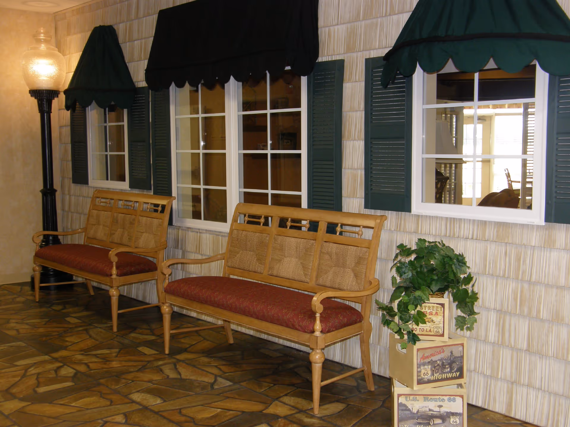 Indoor seating area with two wooden benches against a faux exterior wall with windows and green awnings, a floor lamp, and a potted plant.