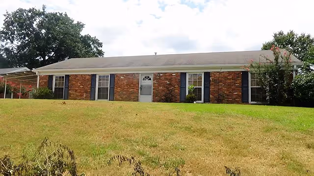 Single-story brick residential building with a gray shingled roof, white door, and blue shutters on the windows, situated on a grassy hill with trees in the background.