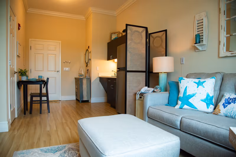A cozy living area in a senior living facility with a gray sofa adorned with blue and white starfish-themed pillows, a matching ottoman, a small table with a lamp and a phone, and a decorative room divider separating the kitchenette. The kitchenette has dark cabinets and a small refrigerator. There is a small dining table with two chairs near a closed door, light wooden flooring, and beige walls with simple decor.