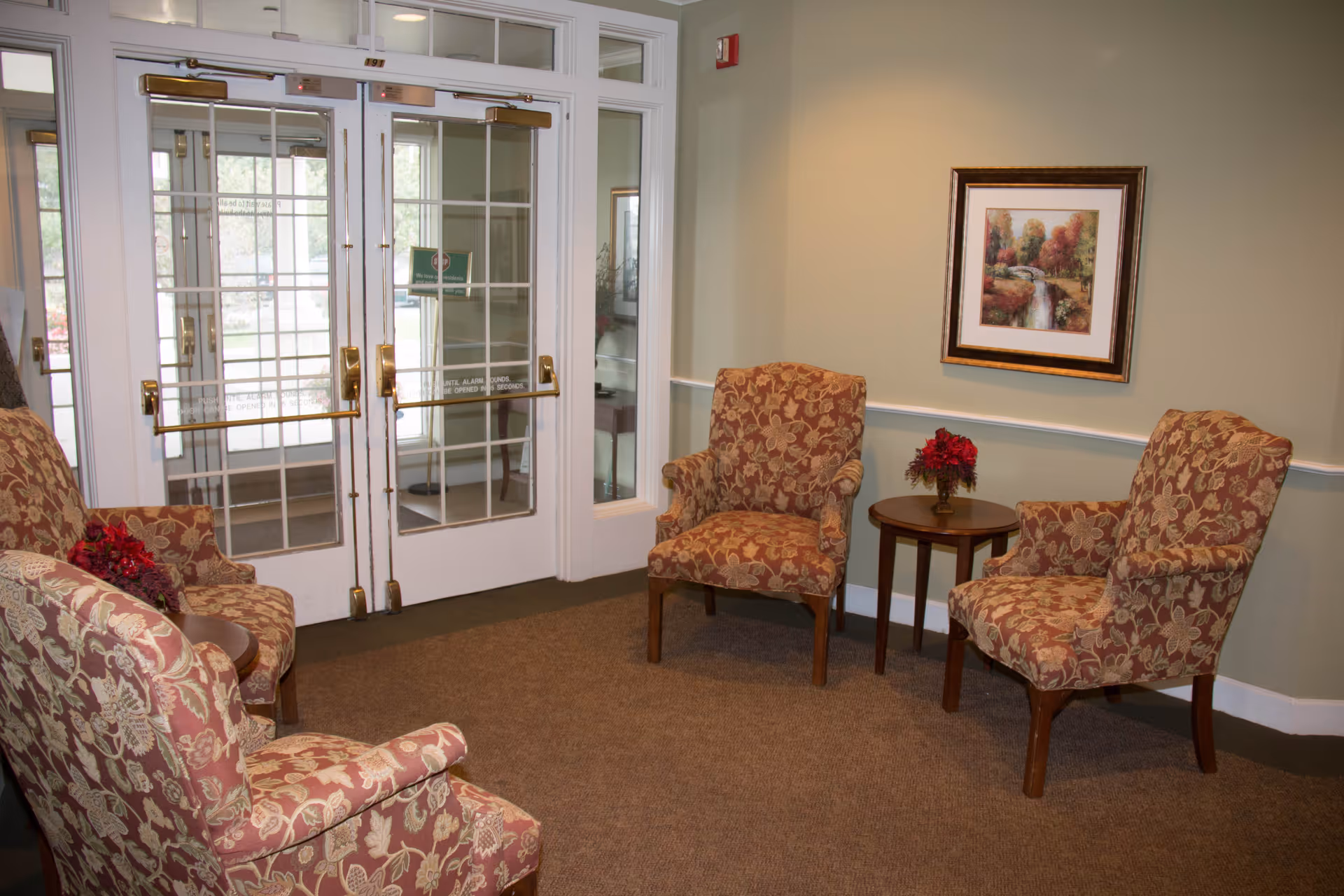 Small lobby seating area with patterned armchairs arranged around a side table, framed artwork on the wall, and glass double doors.
