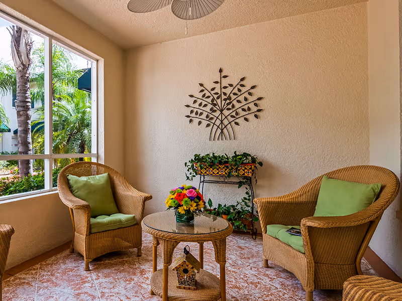 Sunlit seating area with wicker chairs and green cushions, a round wicker table with a floral arrangement, potted plants, and decorative wall art.