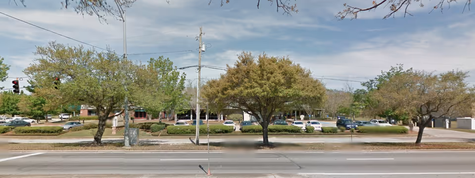 Street-front view of an assisted living building set behind trees and a parking lot across a multi-lane road.