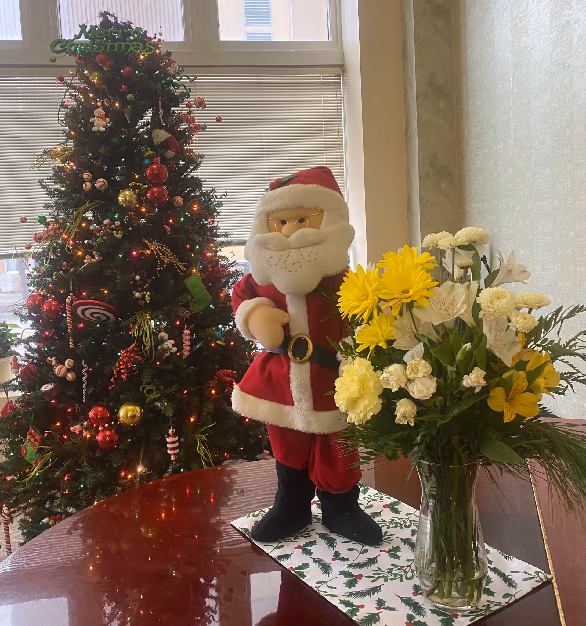 A festive indoor scene featuring a decorated Christmas tree with lights and ornaments, a plush Santa Claus figure dressed in a red suit with white trim, and a glass vase filled with yellow and white flowers on a polished wooden table with a holiday-themed runner.