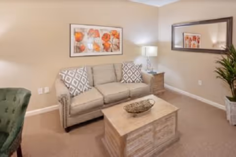 Cozy living room with a beige sofa, wooden coffee table, side table with lamp, wall art, mirror, and a potted plant.