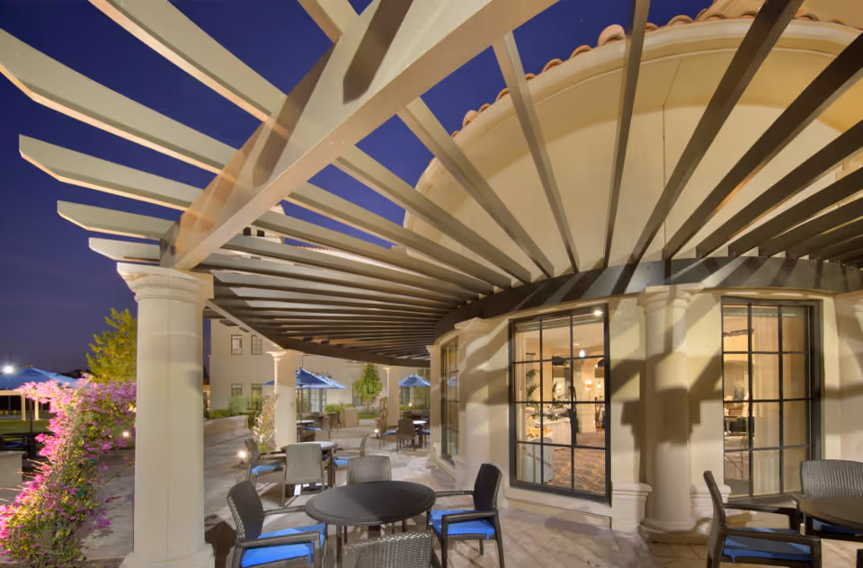 Outdoor patio area at dusk with round tables and chairs under a pergola attached to a building with large windows. There are plants and umbrellas in the background.