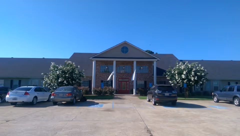 Front exterior view of The Columns of Bossier City facility showing a two-story building with a brick facade, white columns, and a red door entrance. There are several cars parked in front and two flowering trees on either side of the entrance under a clear blue sky.