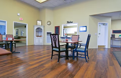 A spacious dining area with wooden flooring, several tables and chairs, light yellow walls, and a serving window labeled 'Kitchen'. There are doors and a white cabinet along the walls, with a clock and framed pictures hanging.