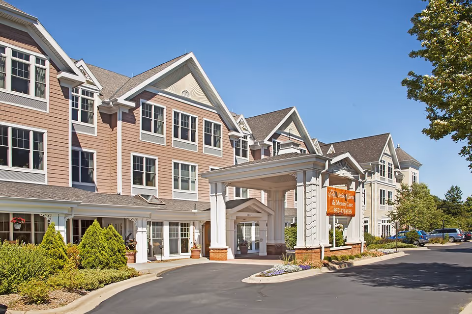 Three-story senior living building with a covered entrance/porte-cochère, many windows, and a landscaped driveway under a clear blue sky.
