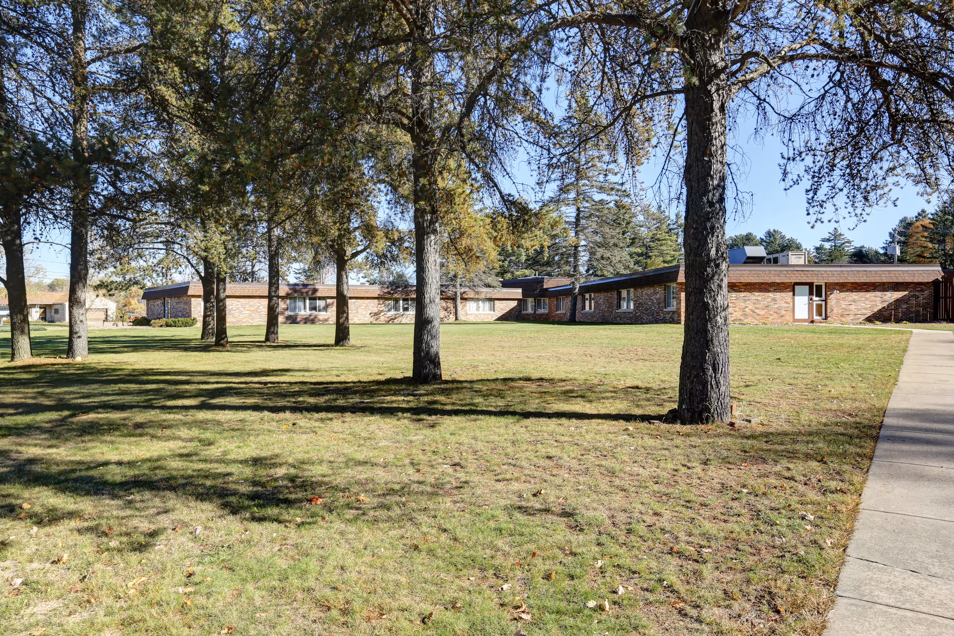 A grassy outdoor area with several tall trees casting shadows on the ground. In the background, there is a single-story brick building with multiple windows and a door. The sky is clear and blue, and a concrete walkway runs along the right side of the image.