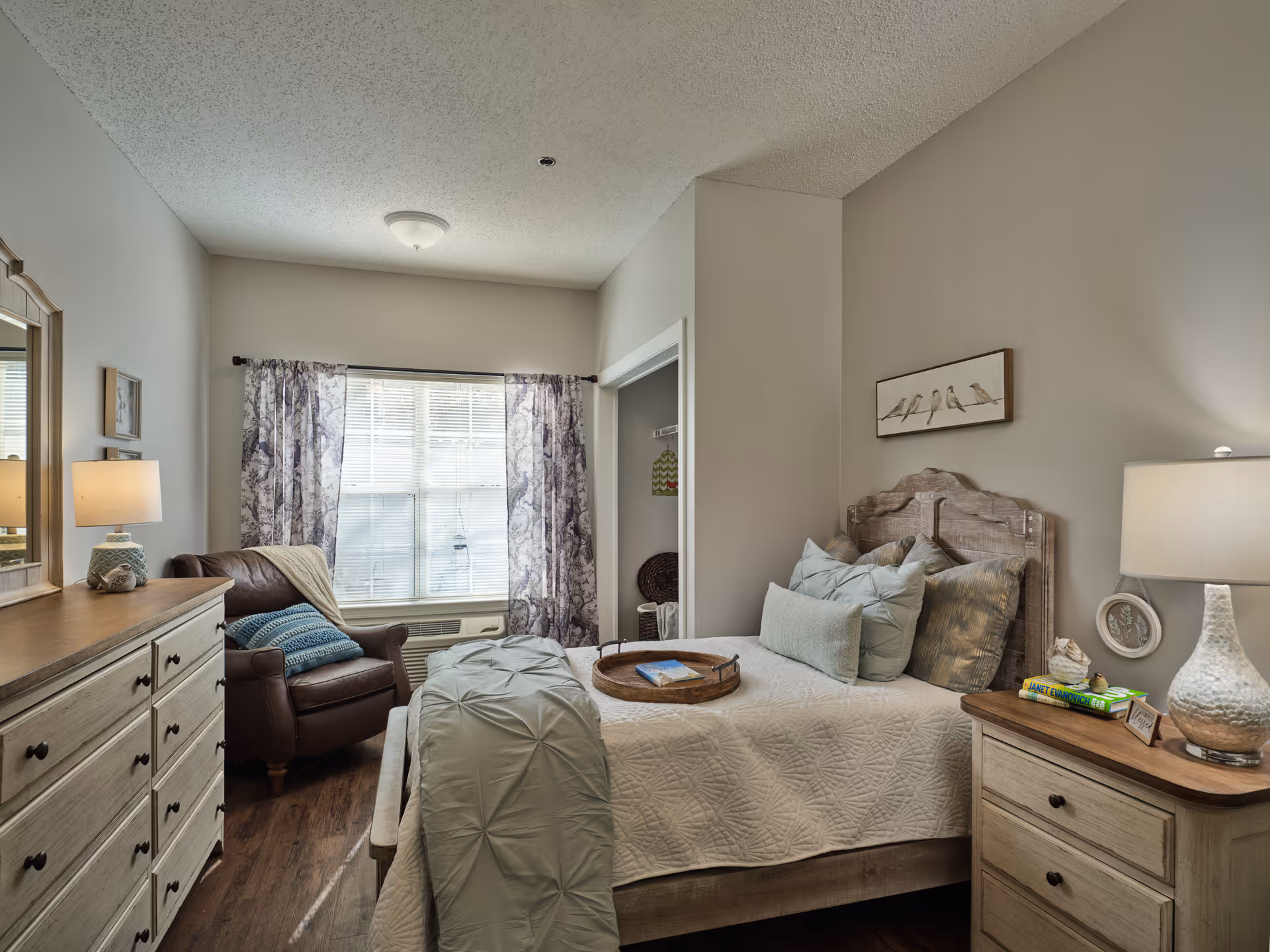 A cozy bedroom in an assisted living facility featuring a bed with multiple pillows and a light quilt, a wooden headboard, a nightstand with a lamp and books, a dresser with a lamp and mirror, a leather armchair with a throw blanket and pillow, and a window with patterned curtains letting in natural light.