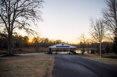 A driveway leads to a covered front entrance of a building at sunset, with bare trees on both sides.