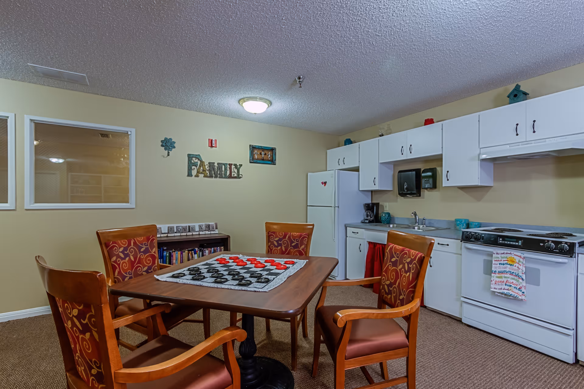 A cozy kitchen and dining area in a senior living facility featuring a wooden table with a checkers game set up, surrounded by four wooden chairs with red patterned cushions. The kitchen has white cabinets, a white refrigerator, a stove with a colorful towel hanging on the handle, a coffee maker, and a sink. The wall is decorated with the word 'FAMILY' and other small decorations. A small bookshelf with books and a sign that says 'I LOVE YOU' is also visible.
