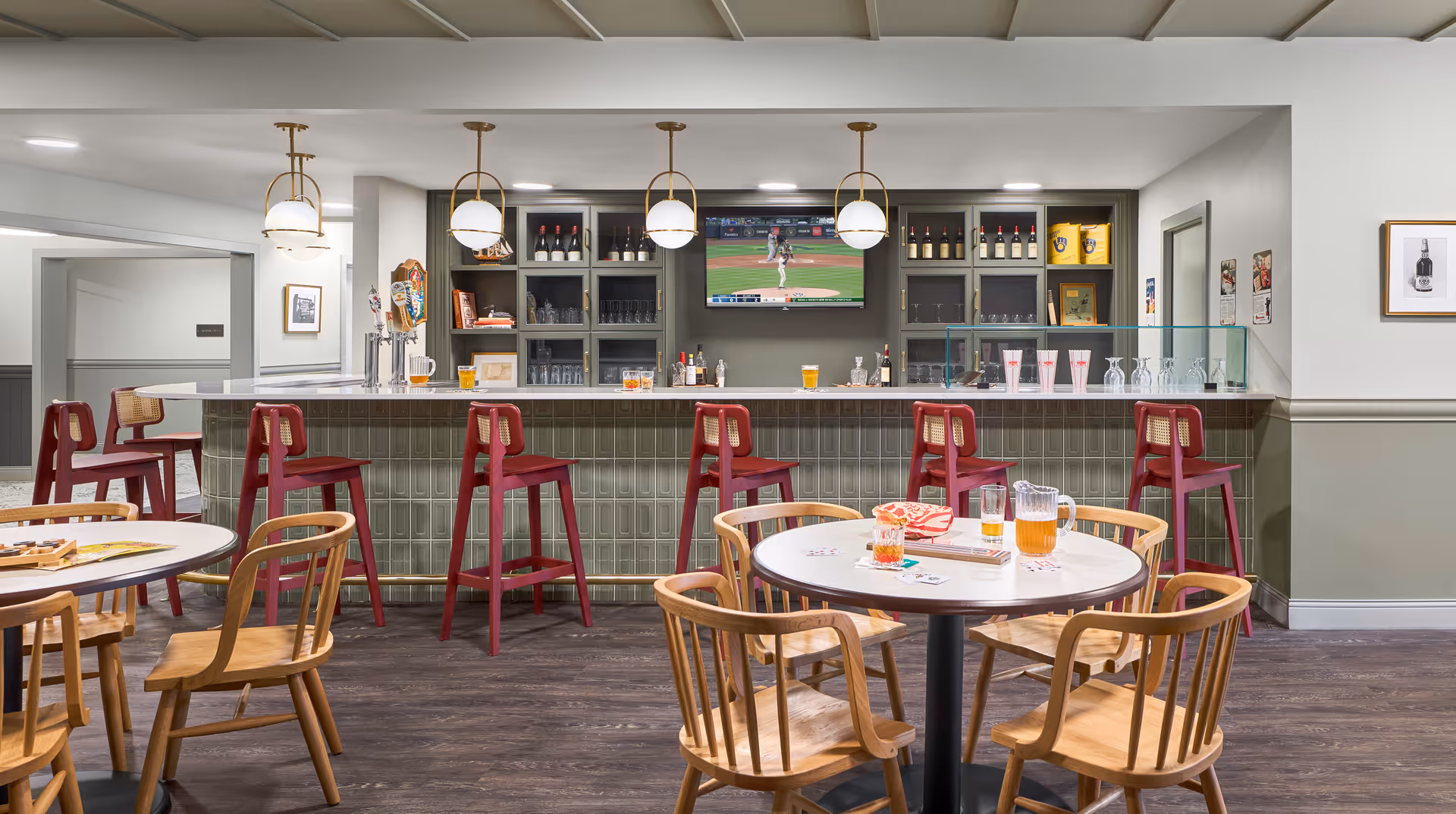 A modern bar area in a senior living facility with a row of red bar stools along a green-tiled counter. Behind the counter are shelves stocked with bottles, glasses, and a television showing a baseball game. In front of the bar are round tables with wooden chairs, some with pitchers and glasses of drinks on them. The room has a clean, bright, and welcoming atmosphere with pendant lights hanging from the ceiling.