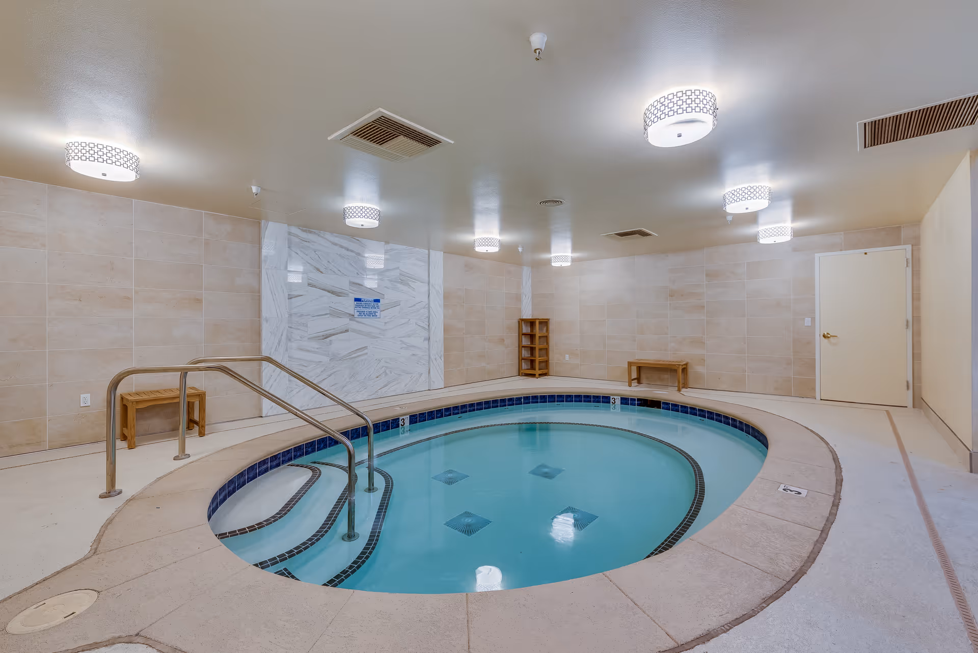 Indoor circular hot tub with metal handrails and steps leading into the water, surrounded by beige tiled walls and floor. The ceiling has multiple modern round light fixtures, and there are wooden benches and a small wooden shelf against the walls.