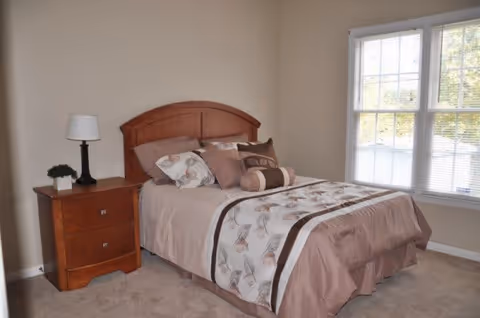 A bedroom with a neatly made bed featuring a floral and brown patterned comforter and multiple pillows. Next to the bed is a wooden nightstand with two drawers, a small potted plant, and a lamp with a white shade. A large window with white blinds allows natural light into the room.