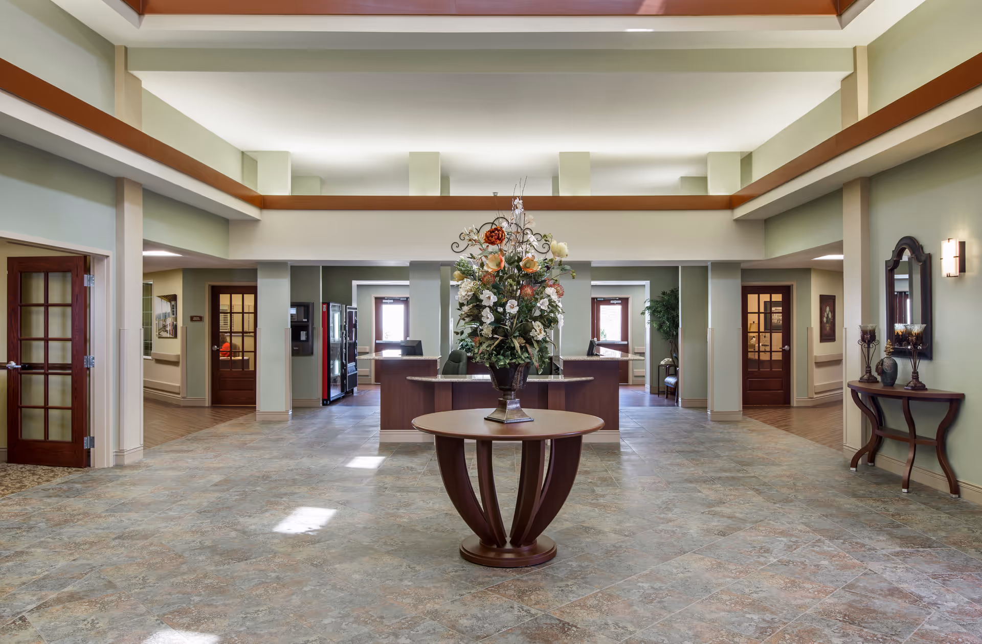 Spacious senior living facility lobby with a round table and large floral arrangement in the center and a reception desk in the background.