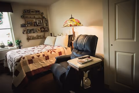 A cozy bedroom with a bed covered by a patchwork quilt, a cushioned recliner chair with a side table holding magazines and a remote control, a Tiffany-style lamp on a nightstand, and shelves on the wall displaying framed photos and small plants near a window.