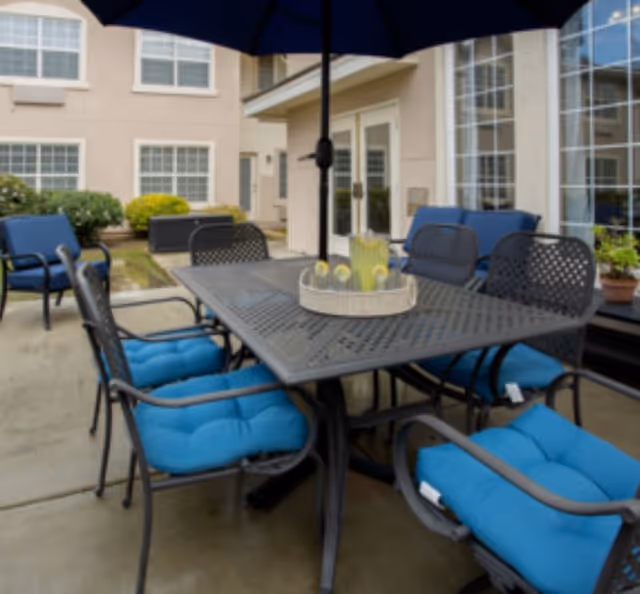 Outdoor patio area with a metal table and six chairs with blue cushions. A large umbrella is positioned in the center of the table, which holds a tray with a pitcher of lemonade and glasses. The patio is adjacent to a building with multiple windows and doors.