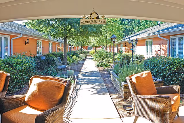 View of a garden pathway lined with bushes and trees, flanked by single-story buildings with orange walls and white trim. Two wicker chairs with orange cushions are visible in the foreground under a covered area. A sign hanging overhead reads 'Welcome To The Garden.'