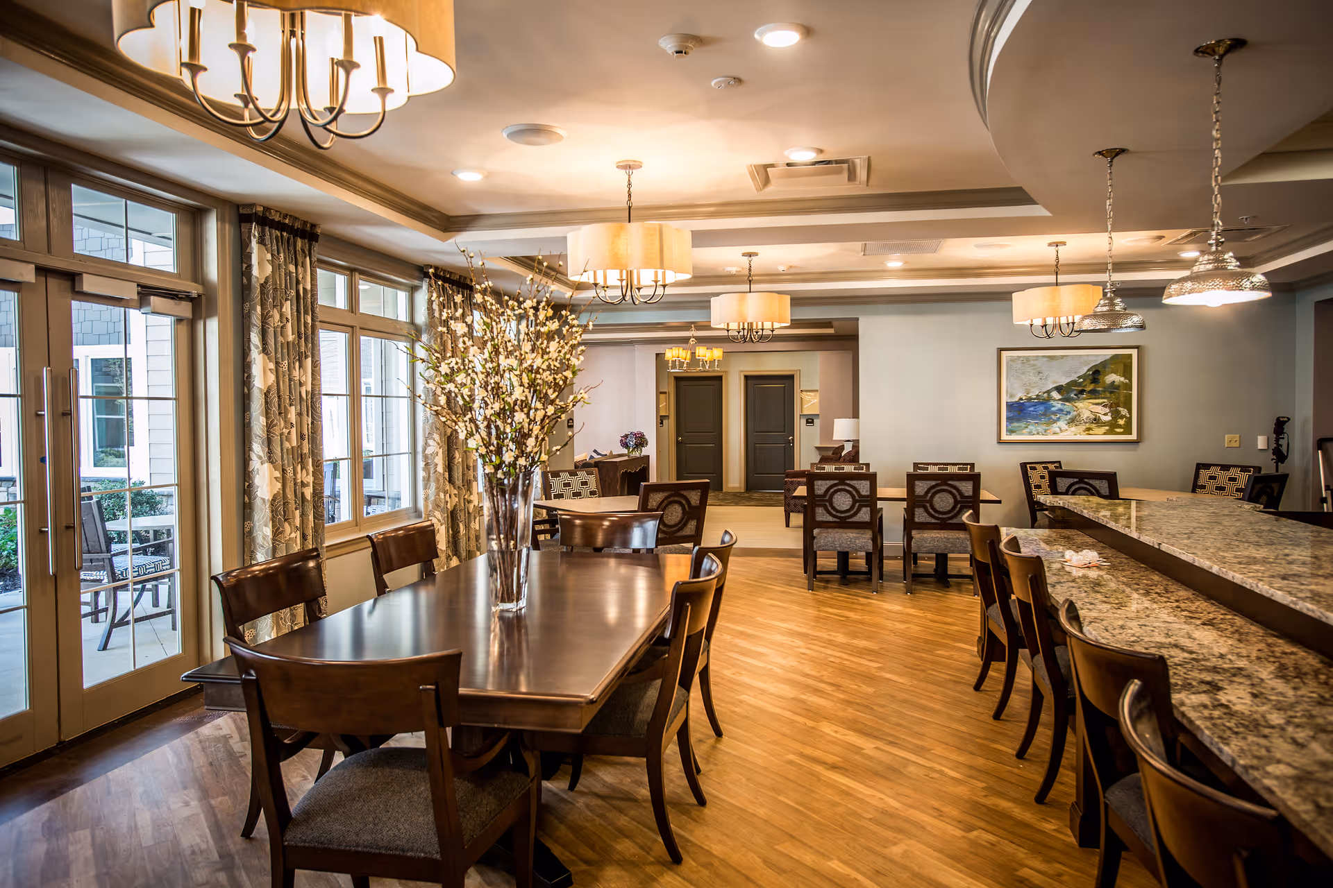 A spacious dining area in a senior living facility with a large wooden dining table surrounded by chairs, a vase with white flowers as centerpiece, multiple chandeliers hanging from the ceiling, large windows with patterned curtains allowing natural light, and a granite countertop bar with stools on the right side.