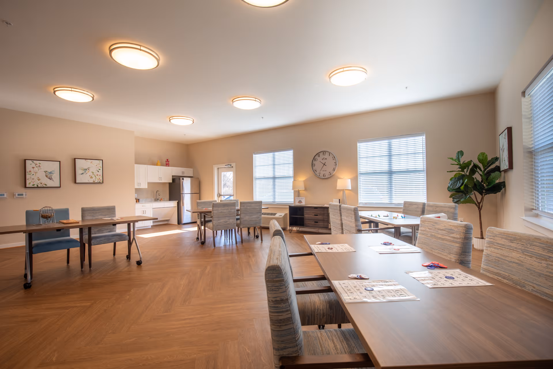 A bright and spacious common room with several tables and chairs arranged for group activities. The room features wood flooring, beige walls, and multiple ceiling lights. There is a small kitchenette with white cabinets and a stainless steel refrigerator in the background. The tables have bingo cards and chips on them, indicating a recreational activity. Two windows with blinds allow natural light to fill the room, and a large clock and two lamps sit on a sideboard against the wall. A potted plant and framed artwork decorate the space.