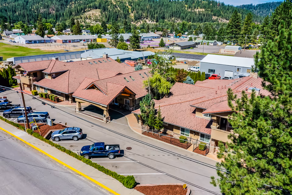 Aerial view of Pinehurst Senior Community showing a large building with a red-tiled roof, surrounded by trees and parked cars along the street. The building has a covered entrance and is situated in a semi-rural area with hills and other buildings in the background.
