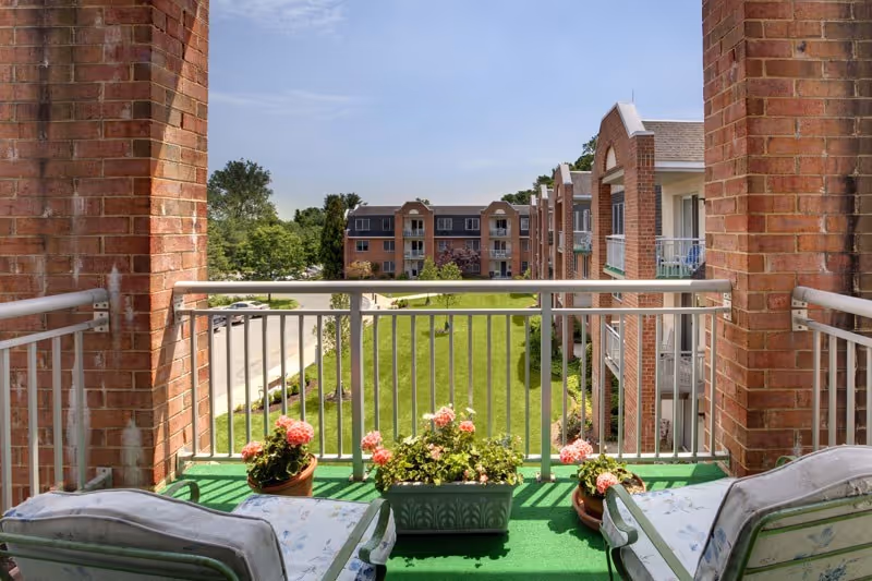 View from a balcony with two cushioned chairs and flower pots with pink flowers on green artificial grass, overlooking a grassy courtyard and a multi-story brick building under a clear blue sky.