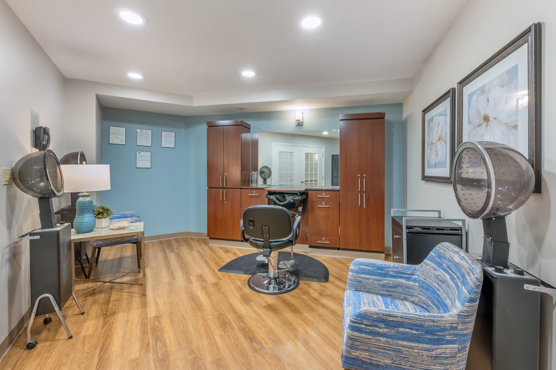 Interior view of a hair salon area in a senior living facility with a salon chair in front of a large mirror and wooden cabinets. There are two hair dryers on stands, a blue patterned armchair, a small table with a lamp and plant, and framed floral artwork on the walls. The floor is wood and the walls are painted light blue and white.