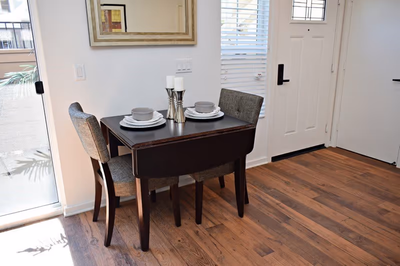 Small dining area with a dark wooden table set for two with gray cushioned chairs, white plates, gray bowls, and silver candle holders. The area is next to a white door and a window with blinds, with wooden flooring and a sliding glass door letting in natural light.