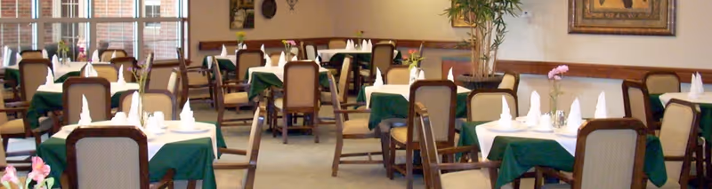 A dining room with multiple tables covered in white and green tablecloths, each set with folded white napkins and small flower arrangements. The room has wooden chairs with cushioned seats and backs, and large windows letting in natural light.
