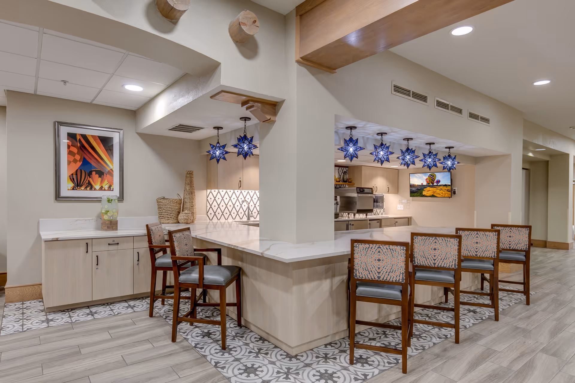 A modern kitchen area in a senior living facility with a long white marble countertop bar surrounded by five wooden chairs with patterned cushions. The kitchen features blue star-shaped pendant lights hanging from the ceiling, stainless steel appliances, and a decorative tile backsplash. On the left side, there is a small counter with a framed picture of hot air balloons and a glass container with infused water. The floor has a mix of wood and patterned tile.