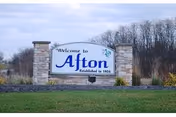 A stone and brick welcome sign for Afton, established in 1854, situated on a grassy area with trees and bushes in the background under a clear sky.