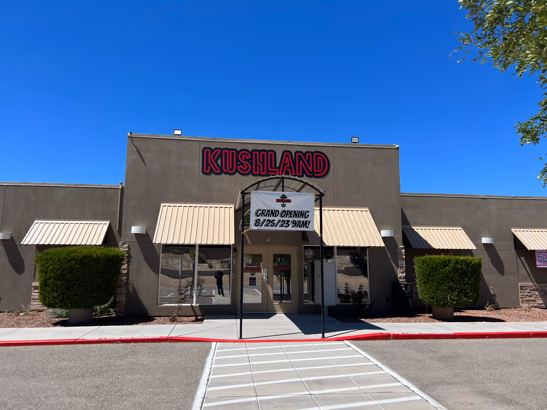 Exterior view of a commercial building with a sign that reads 'KUSHLAND' above the entrance. There is a banner hanging in front of the door announcing a grand opening on 8/25/23 at 9 AM. The building has beige awnings over the windows and doors, with neatly trimmed bushes on either side of the entrance. The sky is clear and blue.