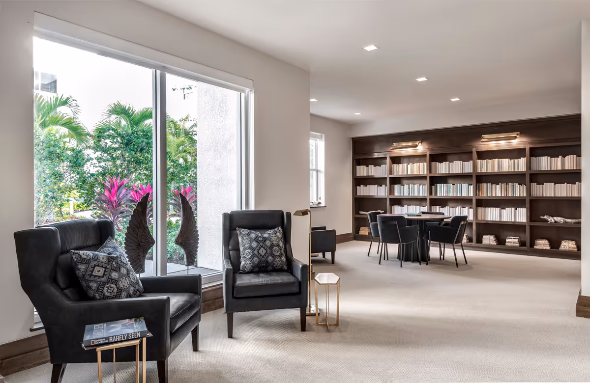 A bright and spacious reading room with large windows showing greenery outside. The room features two black armchairs with patterned cushions near the window, a small side table with a book, and a large wooden bookshelf filled with books along the back wall. There is also a round table with four chairs in front of the bookshelf.
