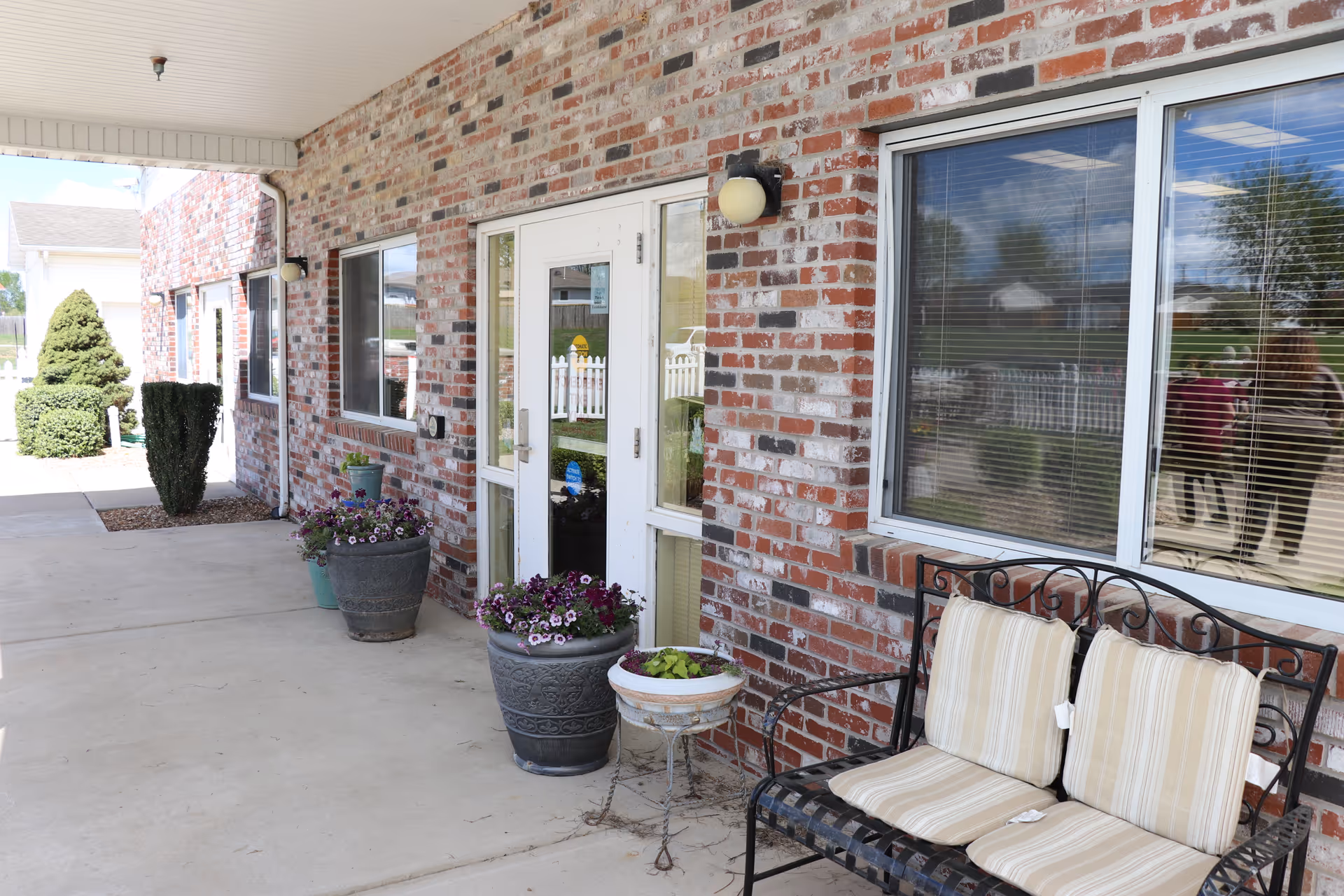 Covered outdoor walkway along a brick building with potted plants and a metal bench with cushions. The building has windows with blinds and a glass door. Reflections of people and outdoor scenery are visible in the window.