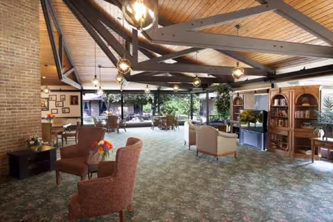 Spacious senior living lounge with seating areas, tables, bookshelves, and warm wood ceiling beams.