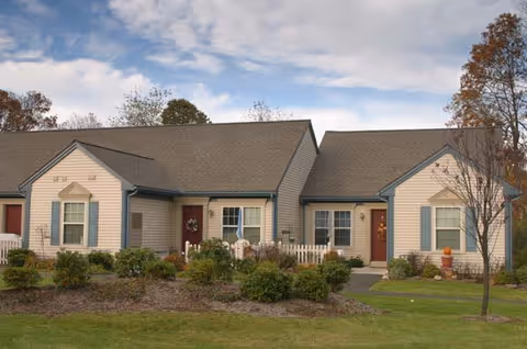 Single-story residential building with beige siding and blue trim, featuring two front doors with wreath decorations, small front porches, landscaped garden beds, and a grassy lawn under a partly cloudy sky.