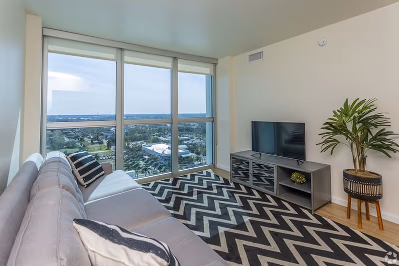 Modern living room with a large gray sofa adorned with black and white pillows, a black and white zigzag patterned rug, a flat-screen TV on a gray media console, a potted plant on a wooden stand, and large floor-to-ceiling windows offering a view of a cityscape and greenery outside.