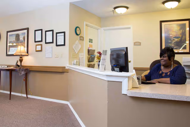 Reception area inside Mercer Terrace facility with a woman sitting behind the front desk working on a computer. The walls are beige with framed pictures and documents, a table with a lamp and some decorations is visible on the left side.