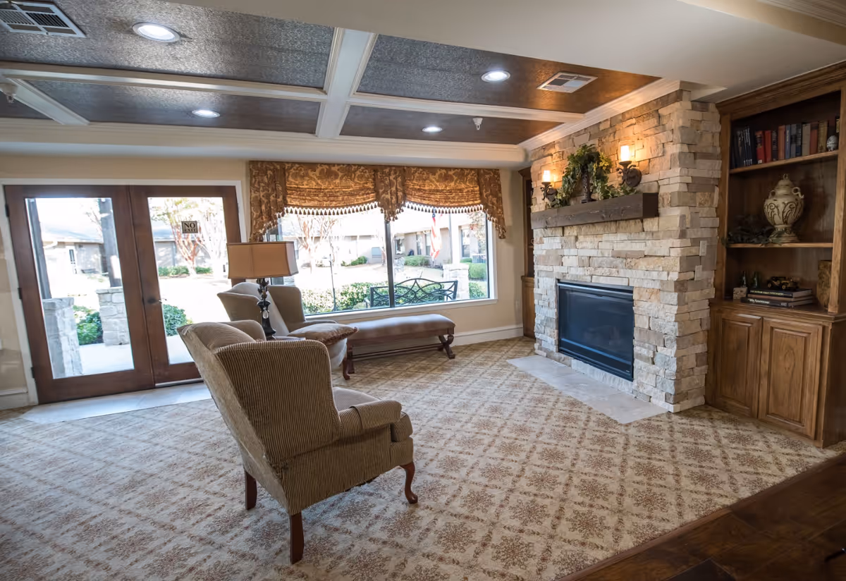 A cozy living room with a patterned carpet, two upholstered armchairs, a cushioned bench near a large window with decorative valances, a stone fireplace with a wooden mantel adorned with greenery and wall sconces, and a built-in wooden bookshelf filled with books and decorative items. Double glass doors lead outside to a garden area.