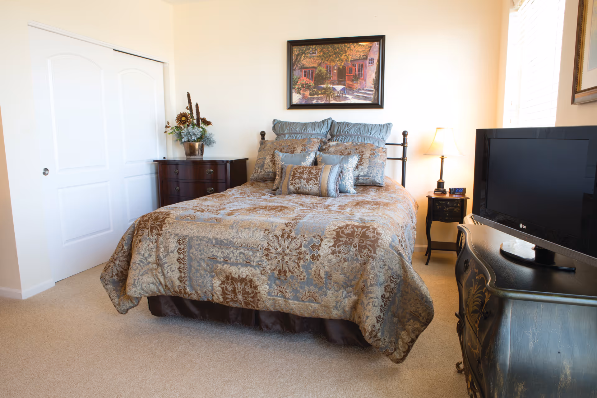 A neatly made bed with a patterned bedspread and multiple pillows in a bedroom. There is a wooden dresser with a floral arrangement on the left side, a nightstand with a lamp and clock on the right side, and a flat-screen TV on a decorative stand in the foreground. A framed painting hangs on the wall above the bed, and there is a window with blinds letting in natural light.