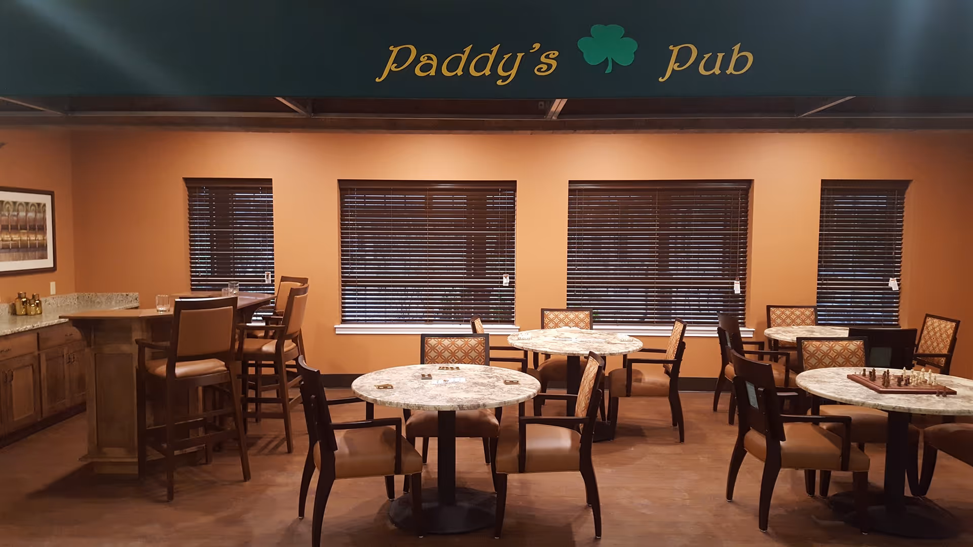 Interior view of a room named Paddy's Pub with several round tables and chairs arranged for seating. The room has warm orange walls, four windows with closed blinds, a countertop with bar stools on the left side, and a chessboard on one of the tables.