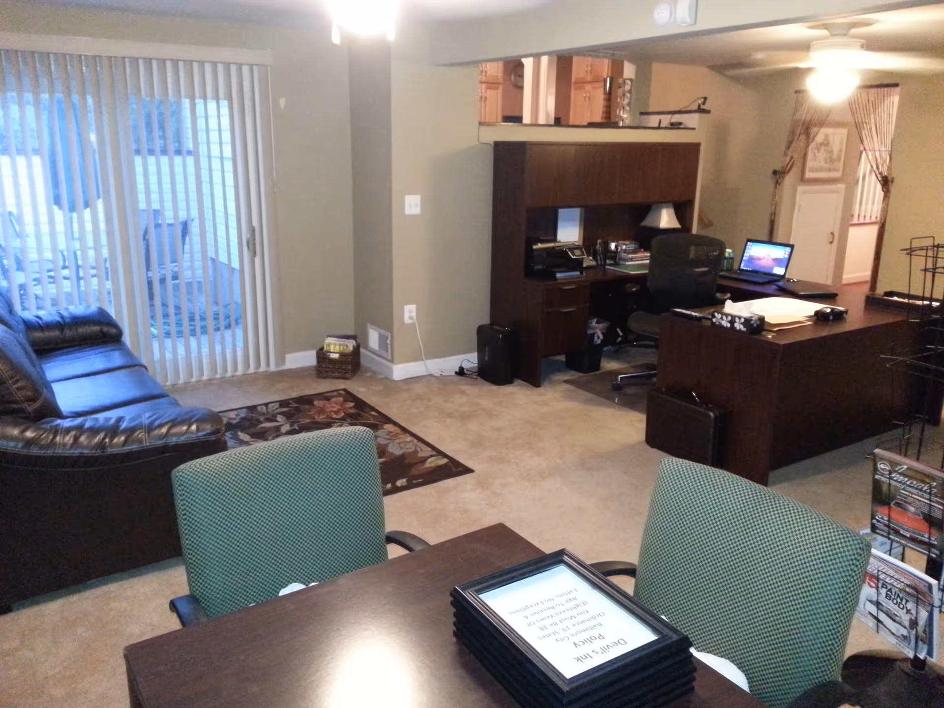 Open living room/office area with a leather couch, sliding glass door, desk with computer, and a table with green chairs.