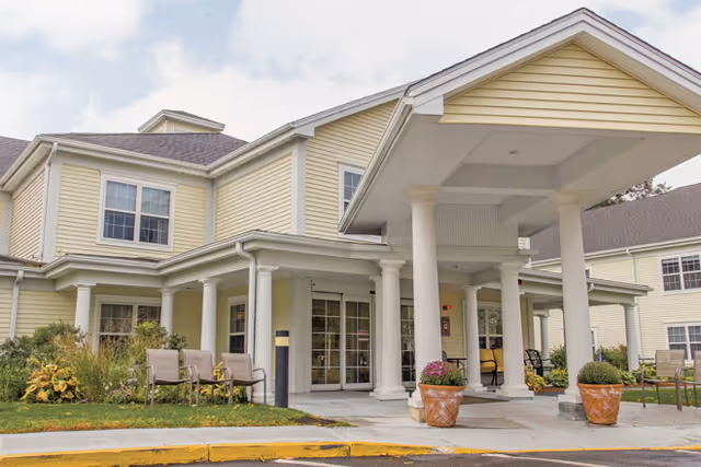 Exterior view of a senior living facility with yellow siding and white columns supporting a covered entrance. There are chairs and potted plants near the entrance, with a small garden area on the left side.