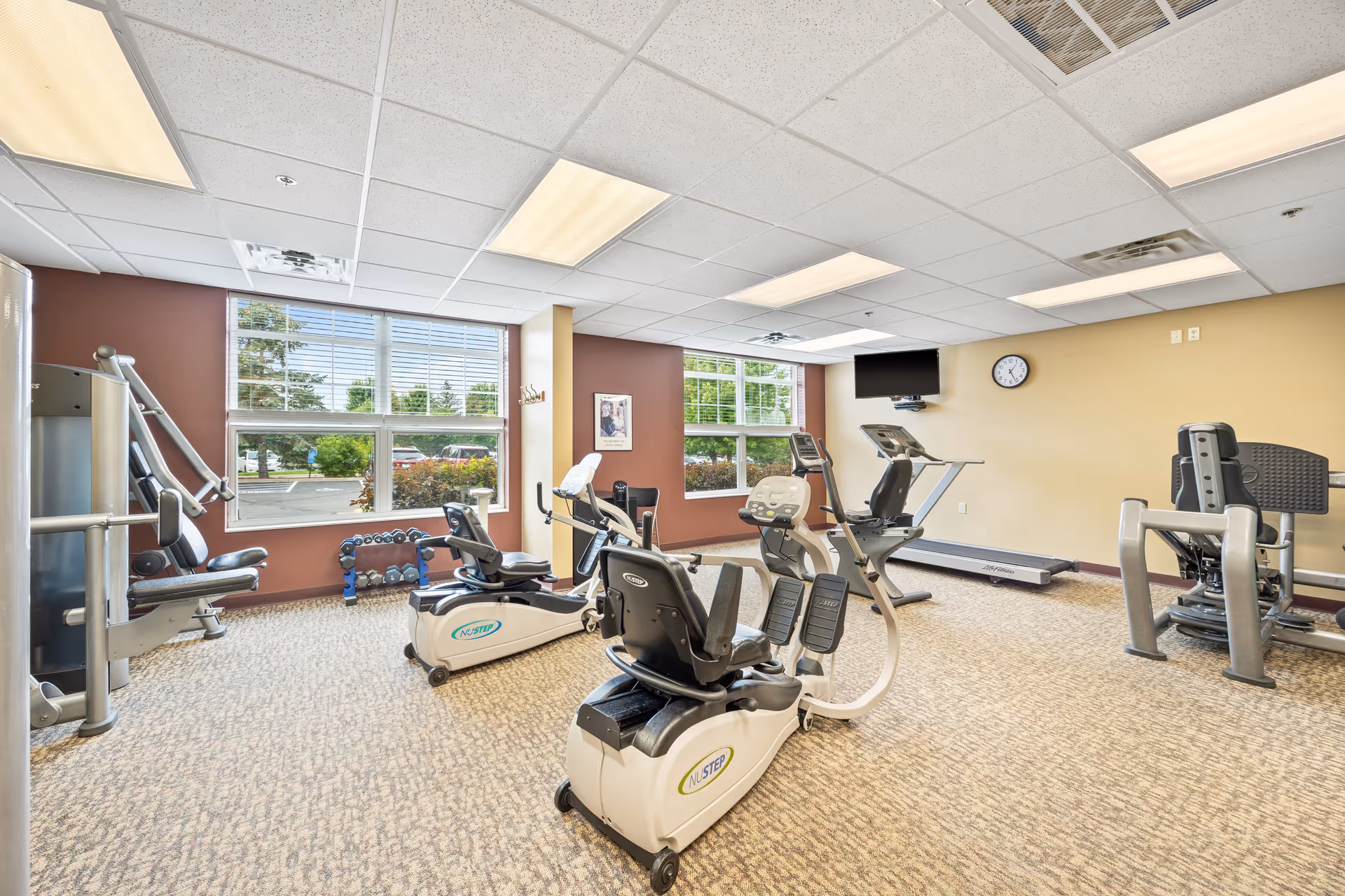 A well-lit exercise room with various fitness equipment including recumbent bikes, a treadmill, weight machines, and dumbbells. Large windows let in natural light and offer a view of greenery outside. A wall clock and a mounted TV are visible on the beige wall.