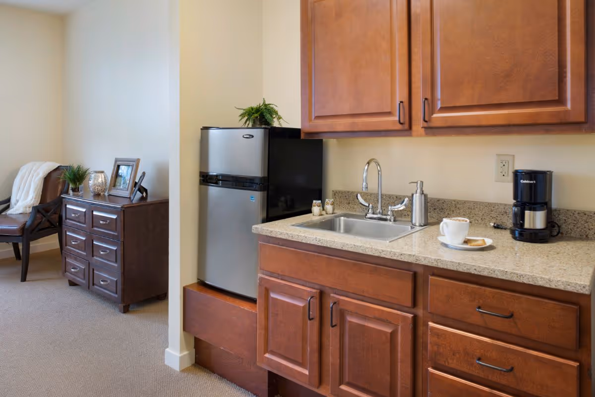 A small kitchenette area with wooden cabinets, a stainless steel mini refrigerator, a sink with a soap dispenser, and a coffee maker on the countertop. To the left, there is a brown leather chair with a white blanket draped over it and a small wooden dresser with decorative items including a framed photo, a small plant, and a candle holder.
