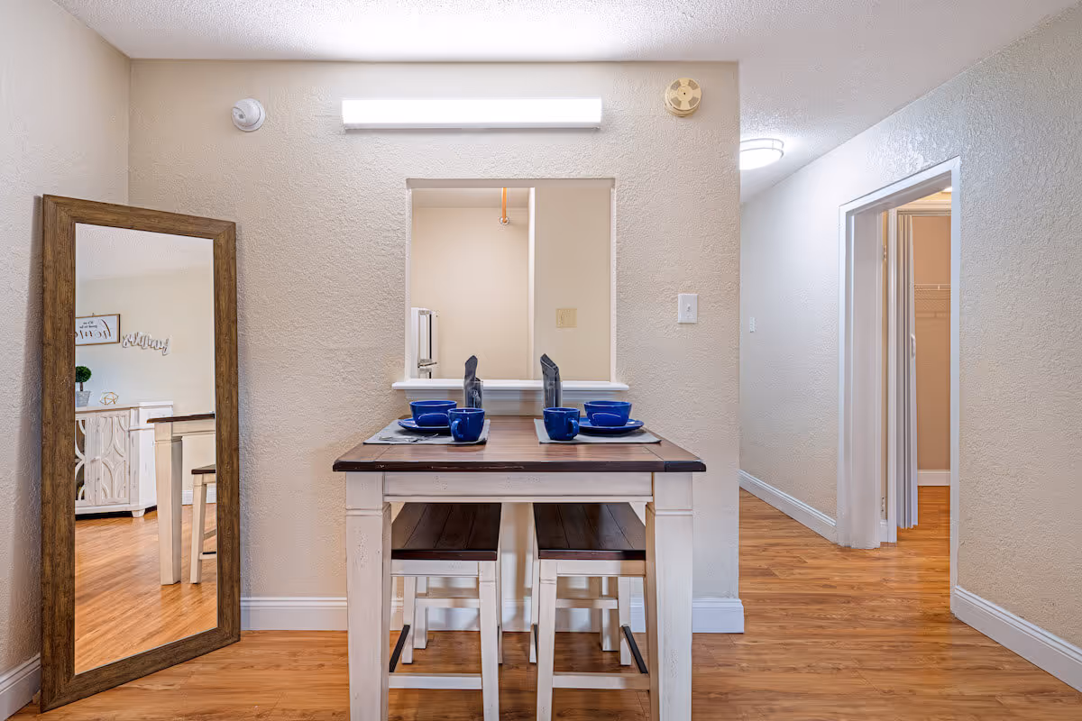 A small dining area with a wooden table set for four with blue dishes and cups. The table has two benches tucked underneath. A large floor mirror with a wooden frame leans against the left wall, reflecting part of a living room with a white cabinet and decor. The walls are beige with a textured finish, and the floor is a light wood laminate. There is a pass-through window above the table opening into the kitchen, and a hallway to the right leads to other rooms.